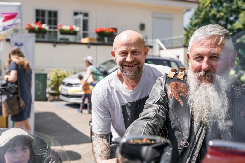 Zwei Männer, einer mit Bart und Lederjacke, der andere mit Glatze, posieren auf einem Motorrad.