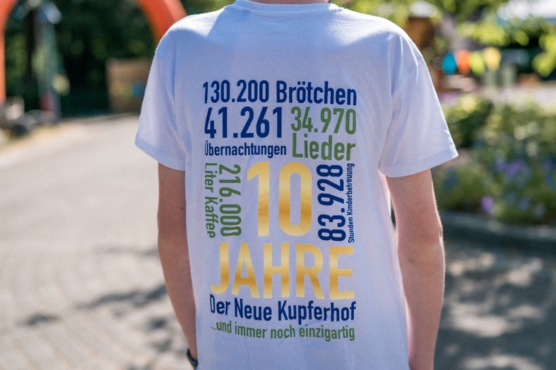 Weißes T-Shirt mit buntem Druck, der Jubiläumszahlen und Informationen enthält.