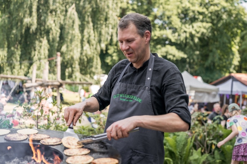 Ein Mann in einem schwarzen Kochoutfit, der Pfannkuchen auf einem Grill im Freien zubereitet.