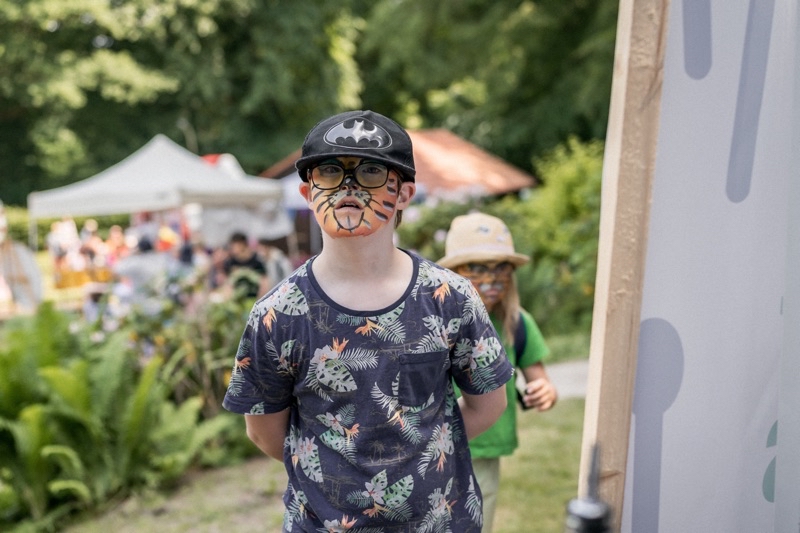 Ein Kind mit Tiger-Gesichtsbemalung und einer Batman-Cap steht im Garten.