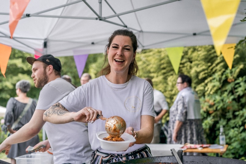 Frau mit langen Haaren in einem weißen T-Shirt, die eine Portion Essen in eine Schüssel serviert.
