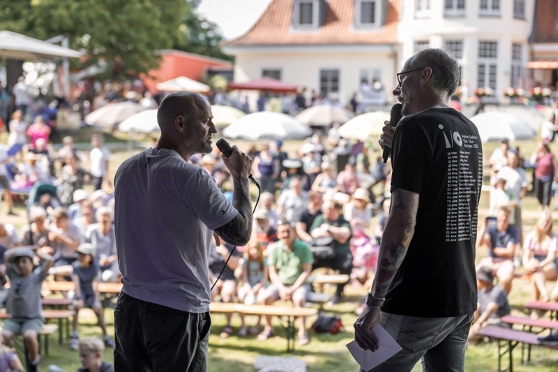 Zwei Männer mit Mikrofonen stehen auf einer Bühne und sprechen zu einem Publikum im Freien.