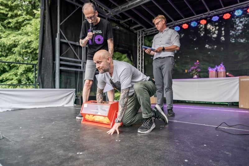 Mann mit Glatze und Tätowierungen sitzt auf einer Bühne, hinter ihm zwei weitere Männer.