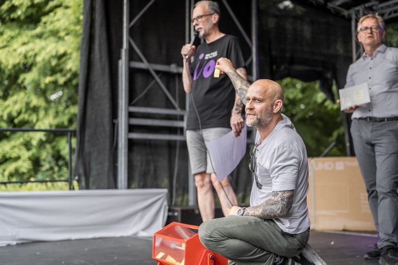 Mann mit Glatze und Tätowierungen sitzt auf einer Bühne, hinter ihm zwei weitere Männer.