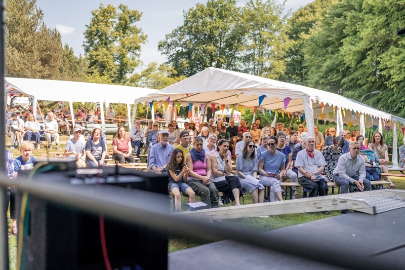 Eine große Menschenmenge sitzt unter Zelten mit bunten Wimpeln in einem Garten.
