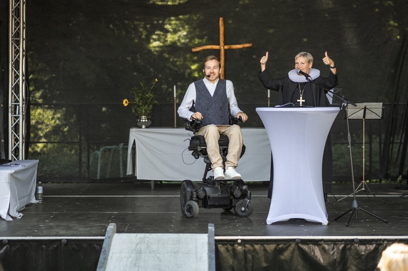 Mann im elektrischen Rollstuhl auf einer Bühne, während eine Pastorin spricht und Daumen hoch zeigt.