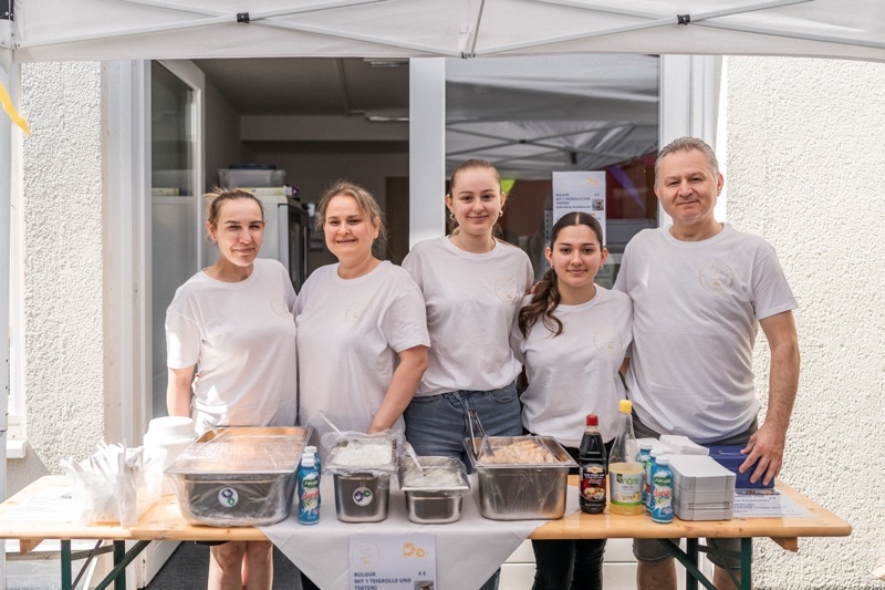 Fünf Personen stehen an einem Essensstand unter einem Zelt mit verschiedenen Speisen und Getränken.