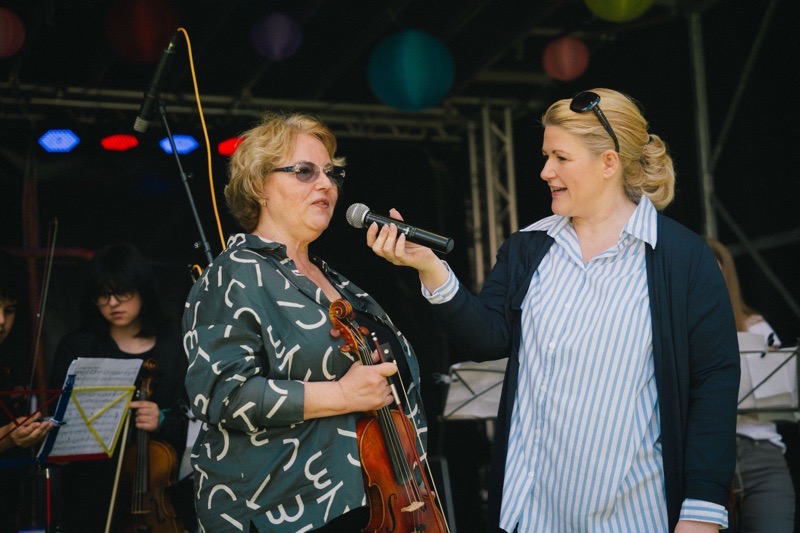 Frau mit Brille hält eine Geige und spricht mit einer anderen Frau, die ein Mikrofon hält.
