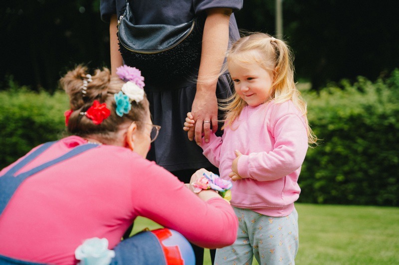 Ein Mädchen in einem rosa Pullover steht neben einer Frau, während eine als Clown verkleidete Frau vor ihr hockt.