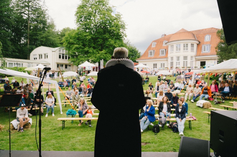 Person mit schwarzem Gewand und Kragen steht vor einer Menge bei einer Veranstaltung im Freien.