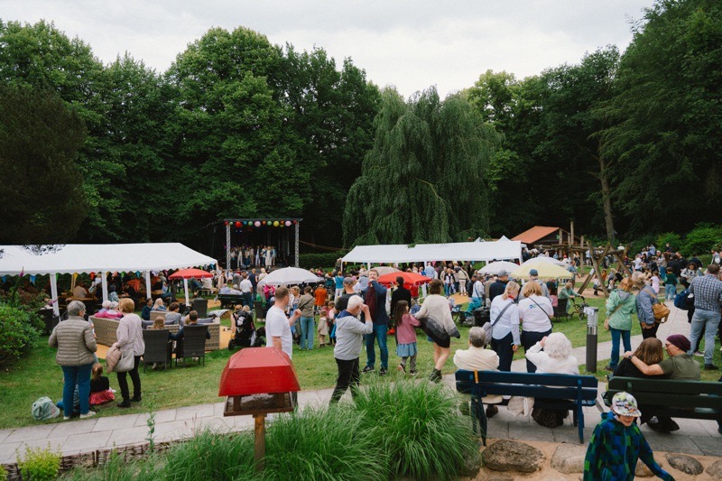 Menschenmenge im Freien mit Zelten und Bäumen im Hintergrund.