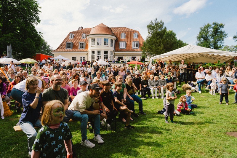 Eine große Menschenmenge sitzt auf einer Wiese während eines Sommerfestes mit Sonnenschirmen und Zelten.