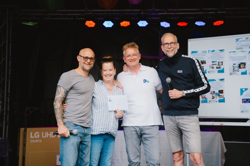 Vier Personen stehen auf einer Bühne und lächeln in die Kamera, zwei davon halten Mikrofone.