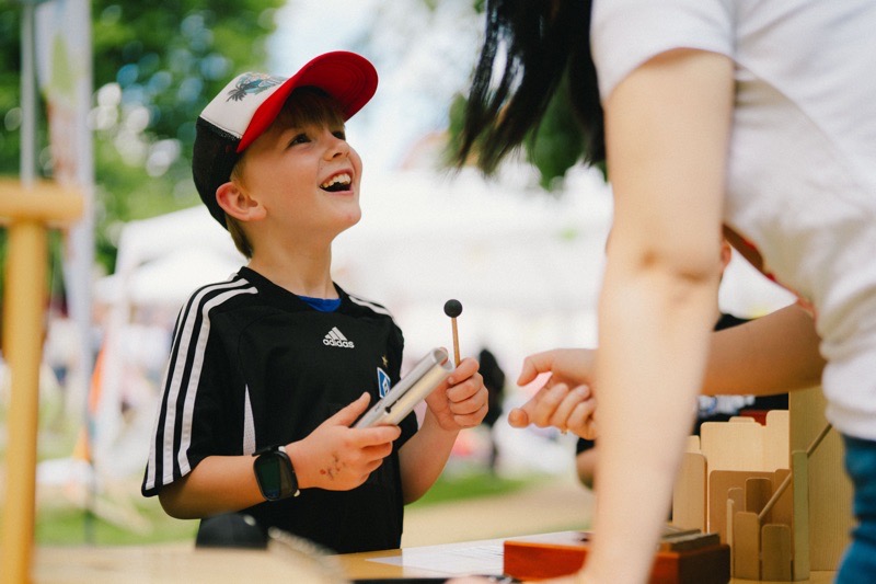 Ein Junge in einem schwarzen Trikot hält ein Musikinstrument und lächelt eine Frau an.