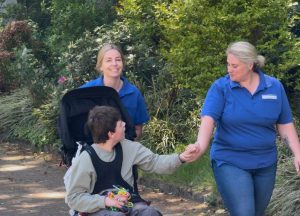 Zwei Pflegekräfte gehen mit einem Kind in einem Rollstuhl und halten seine Hand.