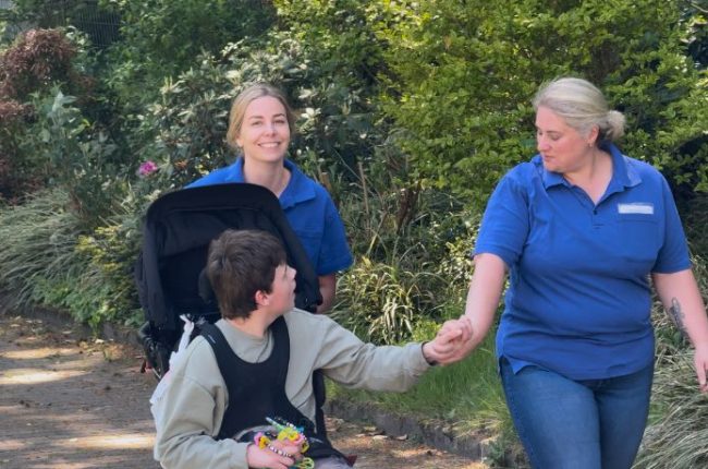 Zwei Pflegekräfte gehen mit einem Kind in einem Rollstuhl und halten seine Hand.