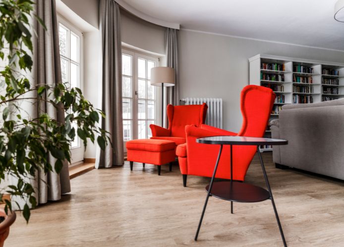 Zwei rote Sessel mit einem kleinen Tisch in einem hellen Wohnzimmer mit Bücherregal im Hintergrund.