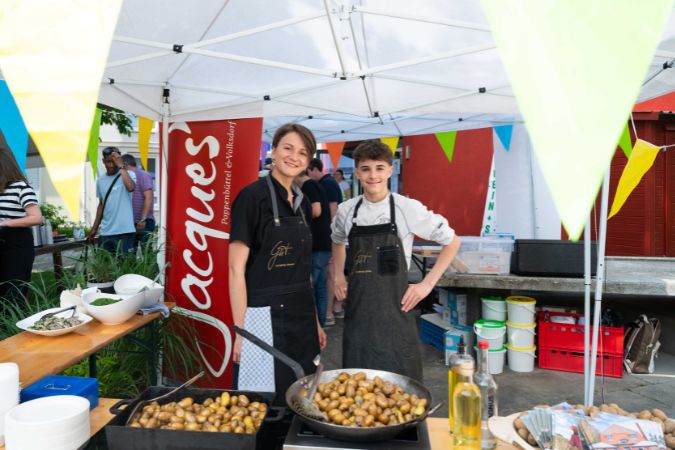 Zwei Personen stehen an einem Kochstand mit einer Pfanne voller Bratkartoffeln auf einem Sommerfest.