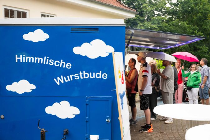 Blauer Imbisswagen mit dem Schriftzug 'Himmlische Wurstbude' und Wolkenmotiven. Viele Menschen stehen an.