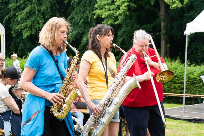 Drei Frauen spielen verschiedene Blasinstrumente, darunter Saxophon und Posaune, während einer Veranstaltung im Freien.