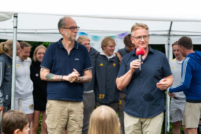 Zwei Männer sprechen auf einer Veranstaltung, während eine Gruppe von Hockey-Spieler: innen im Hintergrund steht.