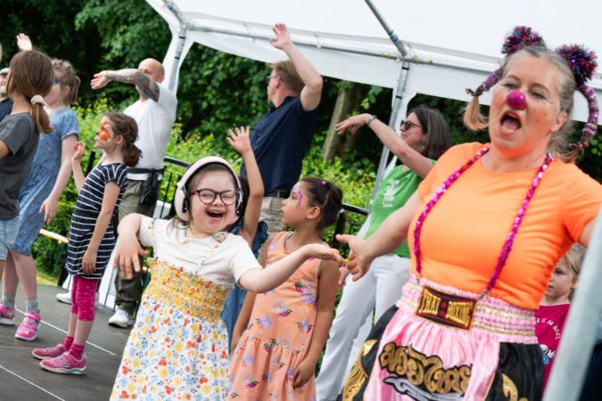 Eine Gruppe von Kindern und Erwachsenen tanzt und lacht auf einer Bühne. Ein Clown ist auch dabei.