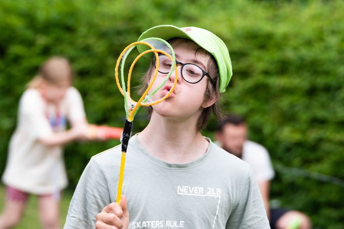 Junge mit Brille und grünem Hut hält einen Seifenblasenstab vor seinem Gesicht.