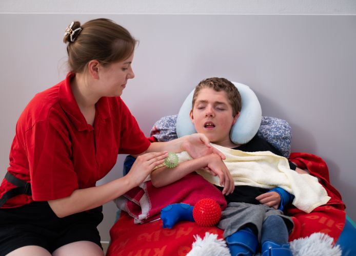 Eine Pflegekraft kümmert sich um einen Jungen, der die Augen geschlossen hat und auf einem Sitzsack mit vielen Decken sitzt.