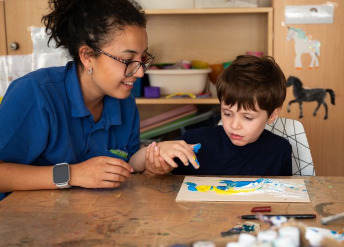 Ein Kind malt mit blauer und gelber Farbe auf einer Holzplatte, während eine Betreuerin ihm hilft.