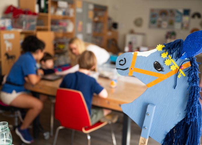 Holzpferdekopf mit blauen und orangefarbenen Details sowie einer blauen Mähne in einem Raum mit Kindern im Hintergrund.
