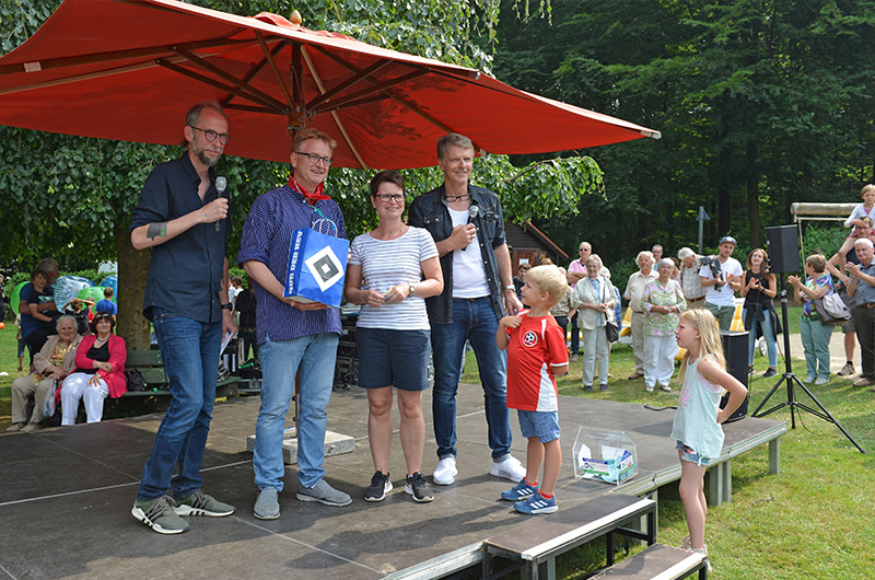 Vier Personen stehen auf einer Bühne während eines Sommerfestes, mit einem großen roten Sonnenschirm im Hintergrund.