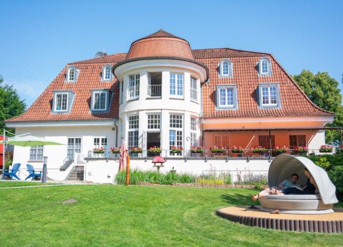 Haus mit runder Erkerfront und rotem Dach, umgeben von einem gepflegten Garten. Ein Mann und eine Frau sitzen in einer Gartenmuschel vor dem Haus.