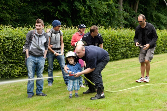 Ein Kind in Feuerwehrkleidung hält einen Schlauch, während ein Erwachsener ihm hilft. Weitere Personen stehen in der Nähe.