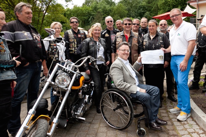 Ein Mann im Rollstuhl und ein stehender Mann halten einen Spendencheck. Hinter ihnen steht eine Gruppe von Motorradfahrer:innen und neben ihnen ein Motorrad.