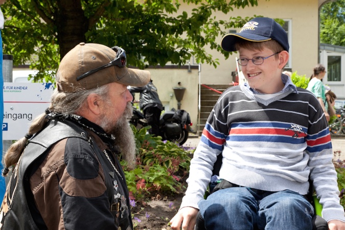 Ein Mann mit Bart spricht mit einem Jungen im Rollstuhl, der ein gestreiftes Shirt trägt.