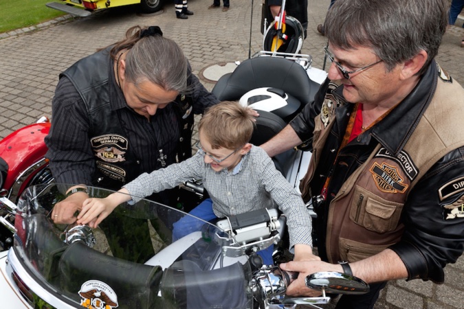 Ein Junge sitzt auf einem Motorrad, während zwei Erwachsene ihm helfen und ihn anlächeln.