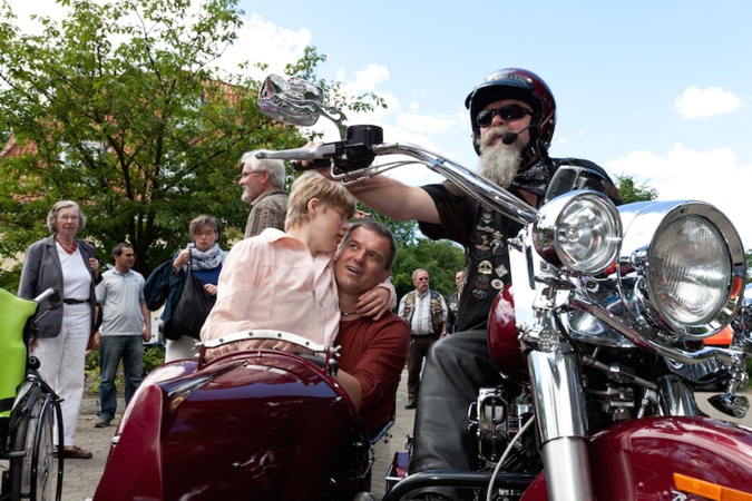 Ein Motorradfahrer mit Bart sitzt auf einem Motorrad. Ein Kind und ein Mann sitzen im Beiwagen neben ihm.