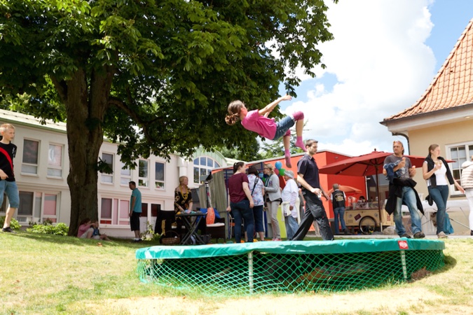 Ein Kind springt auf einem Trampolin, während Menschen im Hintergrund stehen und zuschauen.
