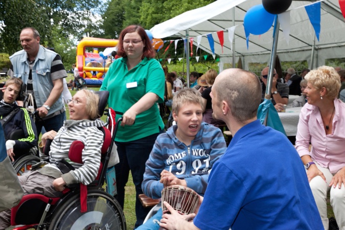 Menschen in Rollstühlen und Helfer:innen bei einem Sommerfest im Freien, mit bunten Fähnchen und einem Zelt.