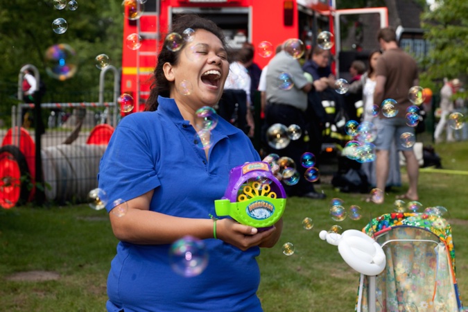 Frau in blauem Polo-Shirt, die Seifenblasen mit einer Maschine erzeugt und dabei lacht.
