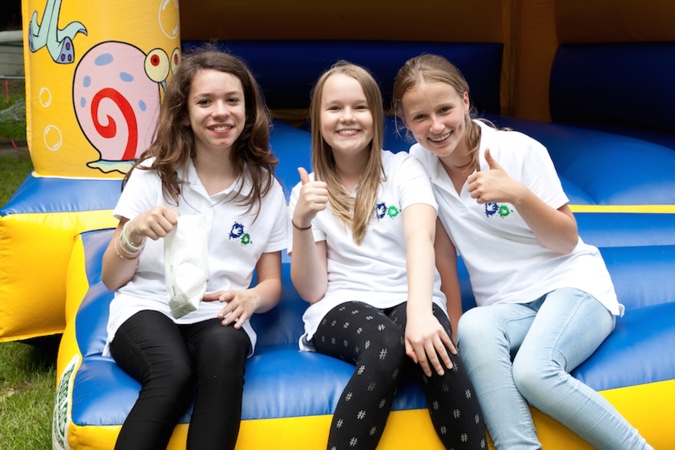 Drei Mädchen in weißen T-Shirts sitzen auf einer Hüpfburg und zeigen Daumen hoch.