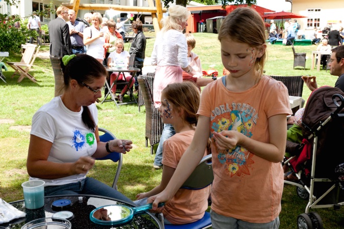 Ein Mädchen hält einen Spiegel, während eine Frau Gesichtsbemalung bei einem anderen Kind macht.