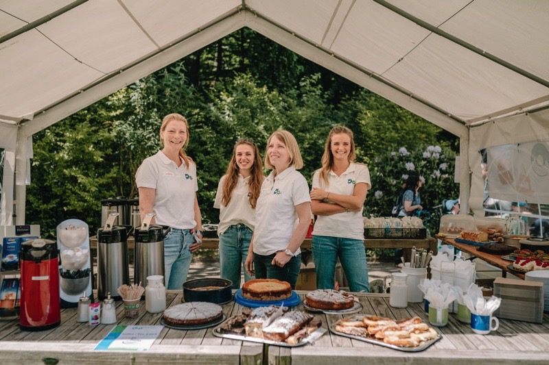 Vier Frauen in weißen T-Shirts stehen an einem Kuchenstand unter einem Zelt.