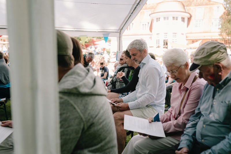 Eine Gruppe von Menschen sitzt in einem Zelt während einer Veranstaltung im Freien und schaut auf ihre Unterlagen.