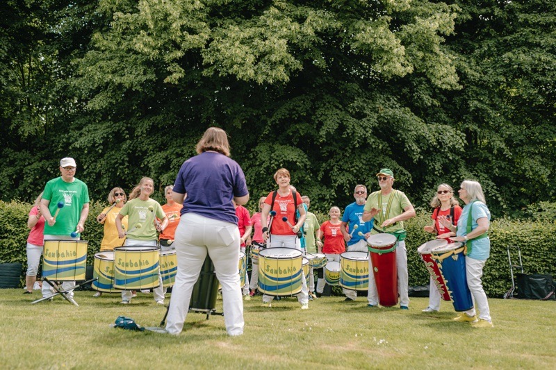 Eine Gruppe von Trommler:innen in bunten T-Shirts spielt auf Trommeln im Freien.
