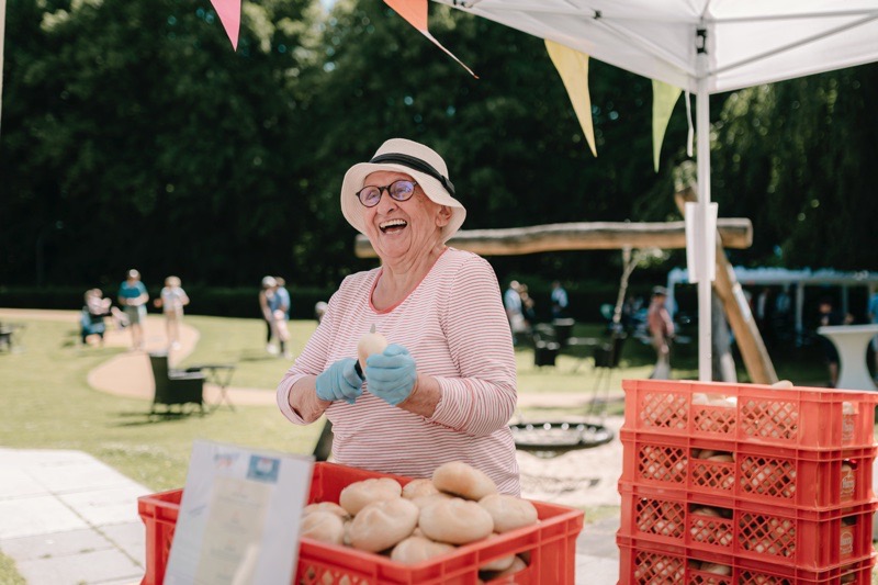 Ältere Frau mit Hut und Handschuhen, die Brötchen an einem Stand beim Sommerfest verkauft.
