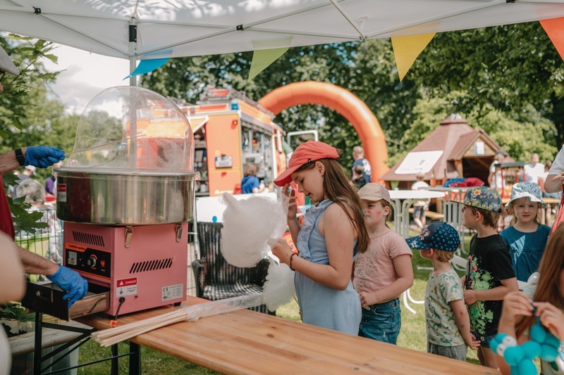 Ein Kind mit einer roten Kappe macht Zuckerwatte an einem Stand mit einer Zuckerwattemaschine. Weitere Kinder stehen an.