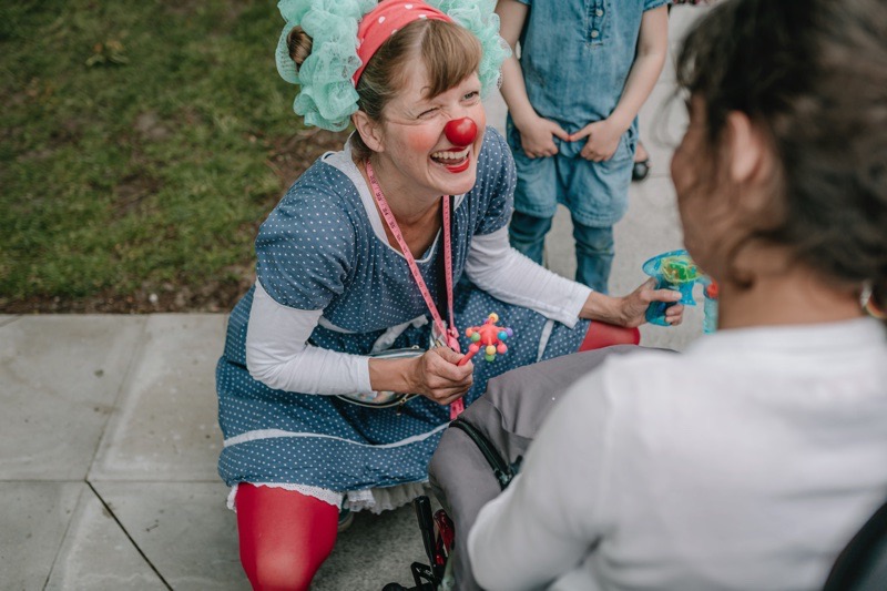Eine Clownin mit roter Nase und buntem Kostüm interagiert mit einem Kind.