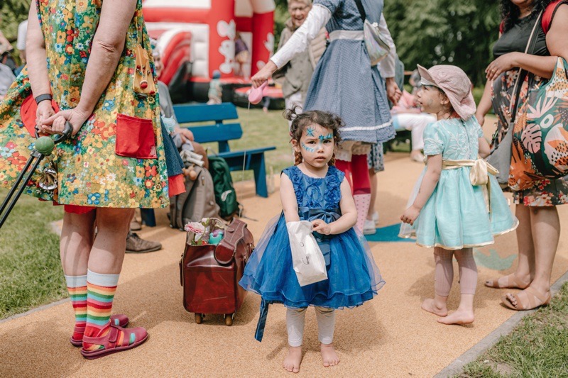 Ein kleines Mädchen in einem blauen Tüllkleid steht barfuß auf einem Weg und schaut in die Kamera. Hinter ihr weitere Menschen.