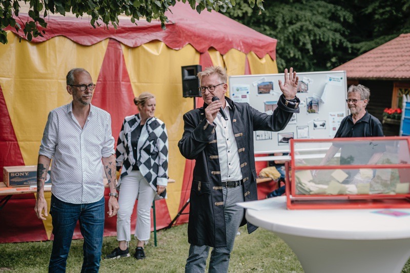 Ein Redner spricht in ein Mikrofon während einer Veranstaltung im Freien, umgeben von Menschen.